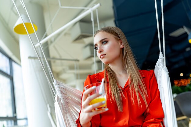 Retrato de uma bela jovem elegante loira sexy no café. Beleza jovem com um copo de suco. Vestido vermelho.