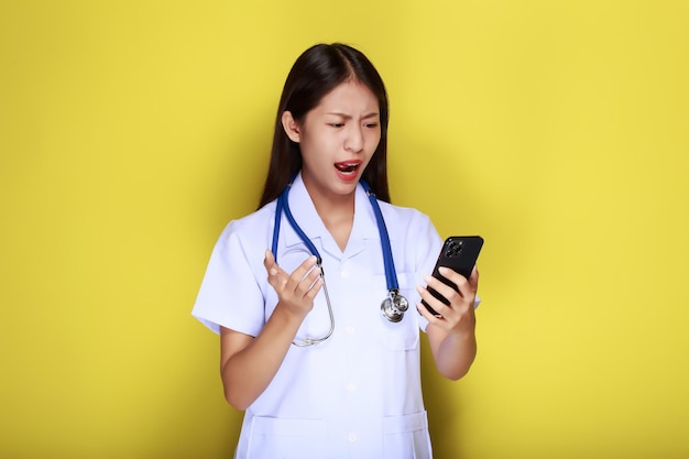 Retrato de uma bela jovem em um fundo amarelo mulher asiática posa com um telefone celular enquanto usa um uniforme de médico e um estetoscópio