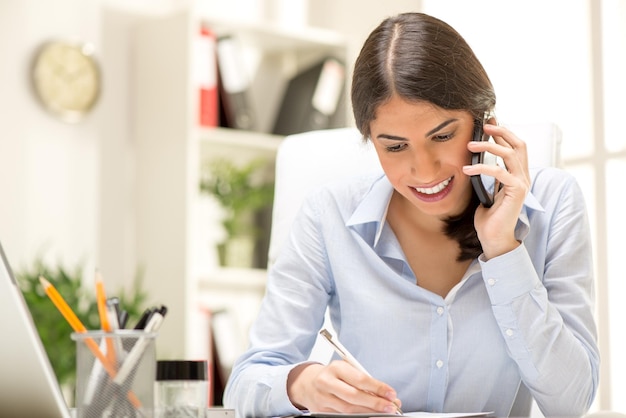 Retrato de uma bela jovem empresária sentada no escritório e telefonando para o celular.