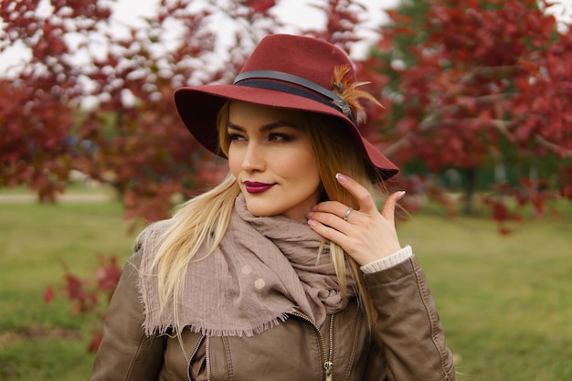 Retrato de uma bela jovem loira elegante com chapéu com pena, lenço e jaqueta, olhando com desprezo para algum lugar distante no parque outono