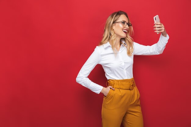 Retrato de uma bela jovem loira em pé isolado sobre um fundo vermelho, tirando uma selfie