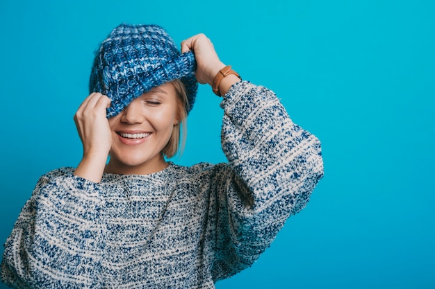 Retrato de uma bela jovem loira rindo com os olhos fechados, enquanto escondia o rosto com um chapéu azul isolado em um fundo azul.