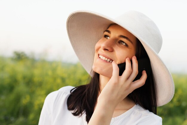 Retrato de uma bela jovem morena atraente com um chapéu falando ao telefone no fundo do campo natural, olhe para cima