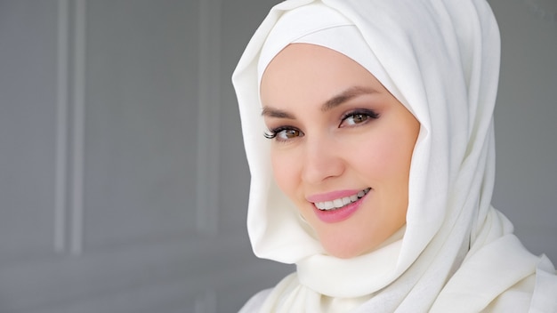 Retrato de uma bela jovem muçulmana árabe usando hijab branco, levantando lentamente os olhos, olhando para a câmera e sorrindo.