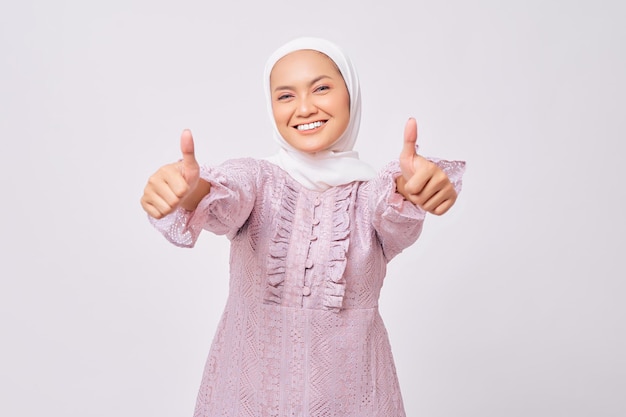 Retrato de uma bela jovem muçulmana asiática sorridente usando hijab e vestido roxo mostrando os polegares para cima gesto para a câmera com uma expressão satisfeita isolada no fundo branco do estúdio