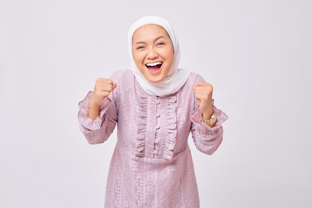 Retrato de uma bela jovem muçulmana asiática usando hijab e vestido roxo levantando os punhos e comemorando a vitória no Ramadã isolada no fundo branco do estúdio