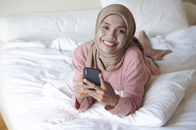 Retrato de uma bela jovem muçulmana asiática usando hijab na cama segurando o celular