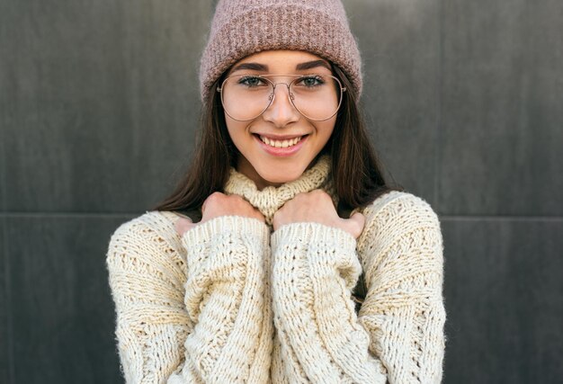 Retrato de uma bela jovem sorridente posando na rua da cidade contra a parede cinza usando suéter rosa, chapéu e óculos transparentes Aluna bonita indo para a faculdade