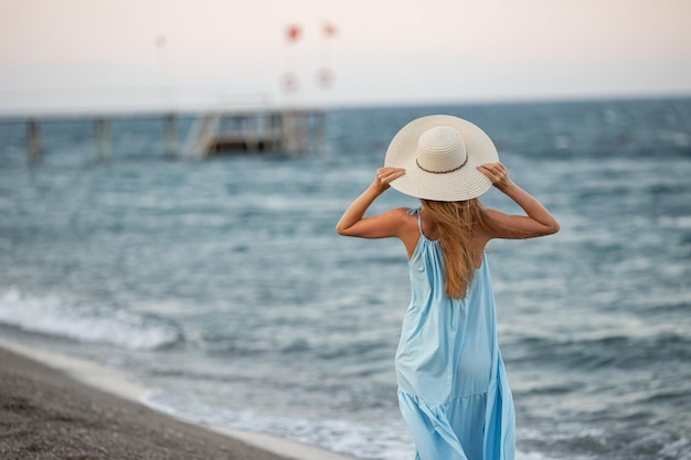 Retrato de uma bela jovem sorridente usando chapéu de palha na praia com o mar ao fundo Menina da moda de beleza olhando para a câmera à beira-mar Mulher bronzeada despreocupada andando na areia e rindo