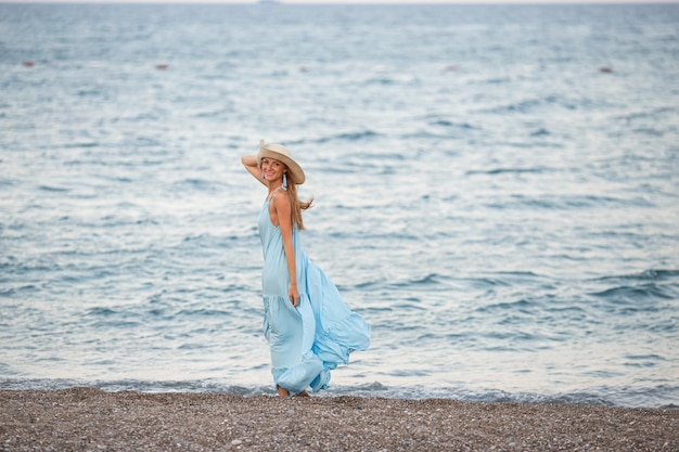 Retrato de uma bela jovem sorridente usando chapéu de palha na praia com o mar ao fundo Menina da moda de beleza olhando para a câmera à beira-mar Mulher bronzeada despreocupada andando na areia e rindo