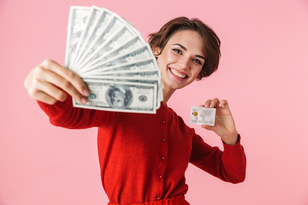 Retrato de uma bela jovem vestindo roupas vermelhas em pé isolado sobre um fundo rosa, mostrando notas de dinheiro, segurando um cartão de crédito