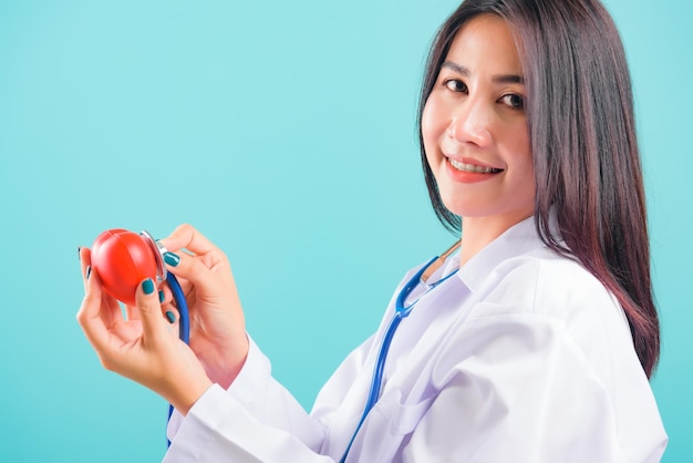 Retrato de uma bela médica asiática sorrindo para ela em pé com estetoscópio examinando um coração vermelho sobre fundo azul, com espaço de cópia para texto