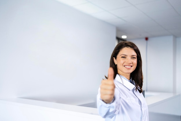 Retrato de uma bela médica sorridente de pé no lobby do hospital e segurando os polegares para cima