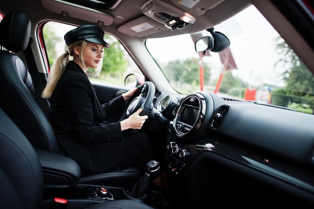 Retrato de uma bela modelo loira sexy de moda em boné e todo preto com maquiagem brilhante, sentar e dirigir o carro vermelho da cidade