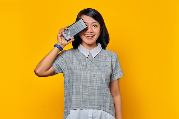 Retrato de uma bela mulher asiática sorridente, cobrindo metade do rosto com smartphone isolado em fundo amarelo