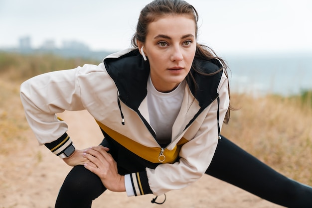 Retrato de uma bela mulher atlética em roupas esportivas, usando fones de ouvido e fazendo exercícios enquanto se exercita na praia