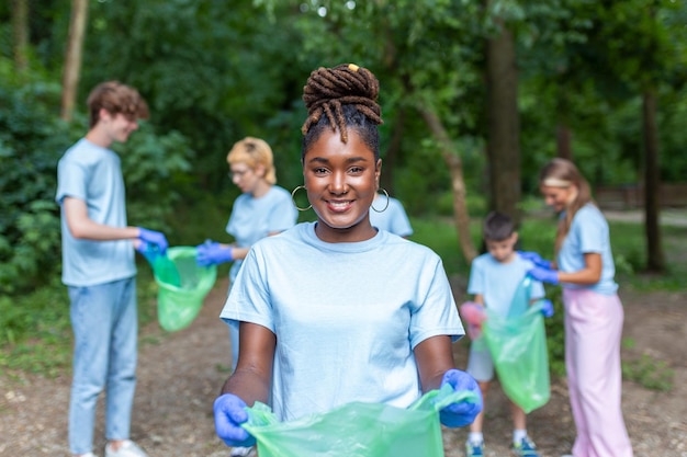 Retrato de uma bela mulher com um pequeno grupo de voluntários no fundo com luvas e sacos de lixo limpando o parque da cidade conceito de preservação ambiental e ecologia Todos vestindo camisas azuis