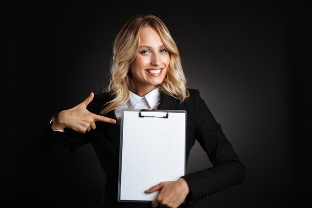 Retrato de uma bela mulher de negócios loira alegre, vestida com roupas formais, em pé isolado sobre a parede preta, mostrando o bloco de notas em branco