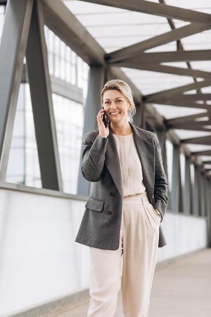 Retrato de uma bela mulher de negócios maduros de terno e jaqueta cinza sorrindo e falando ao telefone em um moderno fundo de edifícios urbanos e de escritórios