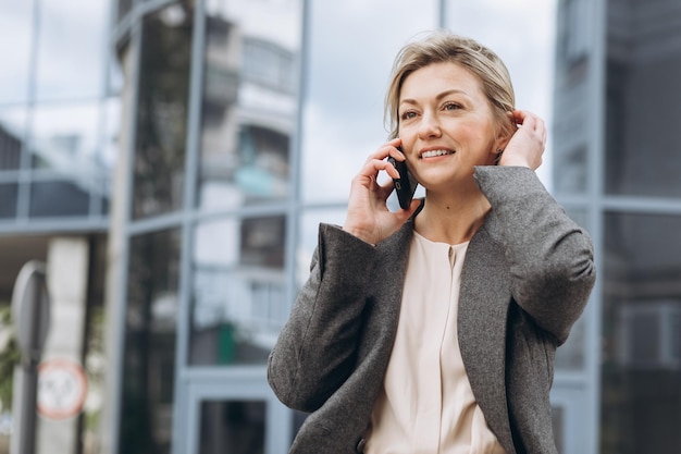 Retrato de uma bela mulher de negócios maduros de terno e jaqueta cinza sorrindo e falando ao telefone sobre o moderno fundo de edifícios urbanos e de escritórios