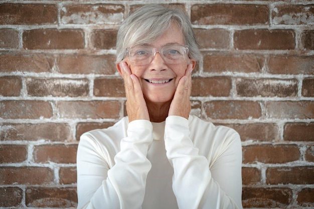 Retrato de uma bela mulher sênior com cabelos brancos e óculos olhando para a câmera sorrindo parede de tijolos no fundo