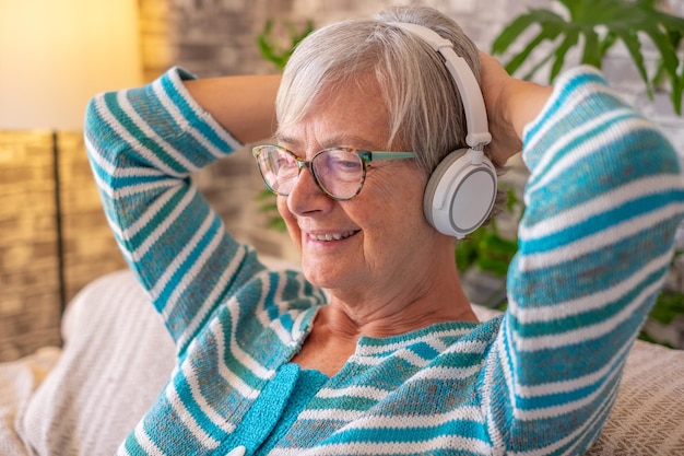 Retrato de uma bela mulher sênior usando fones de ouvido sentado no sofá em casa ouvindo música ou olhando para vídeo sorrindo conceito de senhora relaxada aproveitando a aposentadoria