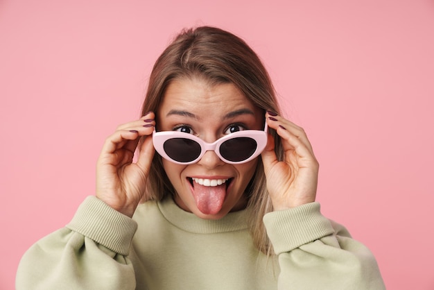 Foto retrato de uma bela mulher sorridente segurando óculos escuros e mostrando a língua isolada na parede rosa