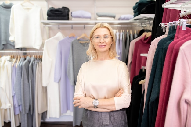 Retrato de uma bela vendedora madura sorrindo para a câmera enquanto está na loja de roupas e espera pelos clientes