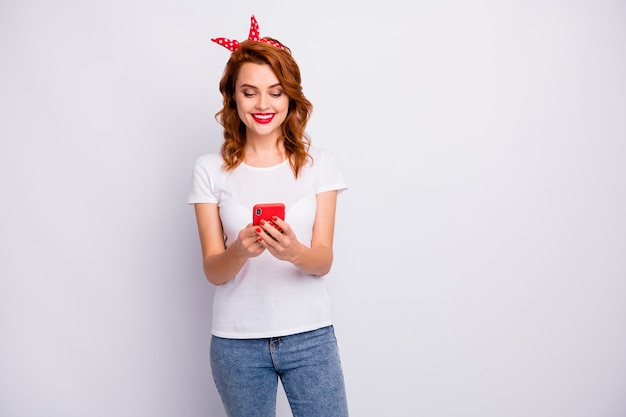 Retrato de uma blogueira positiva e alegre usando o celular, lendo notícias nas redes sociais, vestindo uma camiseta isolada sobre uma parede branca