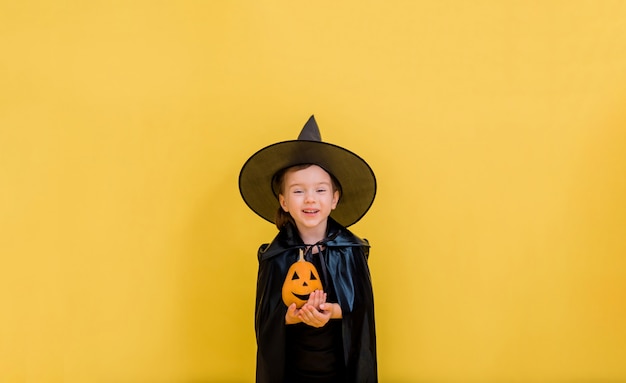 Retrato de uma bruxinha feliz em um chapéu com uma abóbora laranja em um amarelo isolado