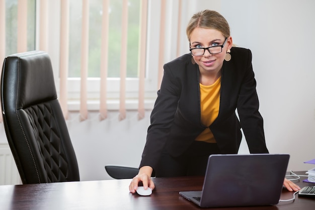 Retrato de uma chefe feminina em uma jaqueta em seu escritório Mulher de negócios está sentada à mesa com laptop