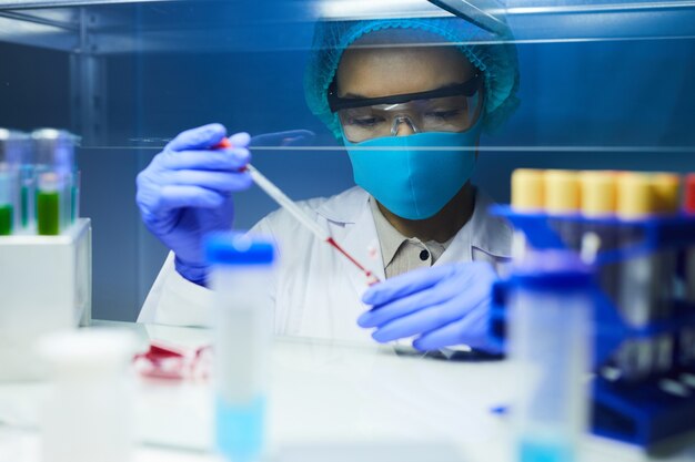 Retrato de uma cientista usando máscara e equipamento de proteção, deixando cair amostras de sangue em tubos de ensaio enquanto trabalhava em pesquisas em laboratório.
