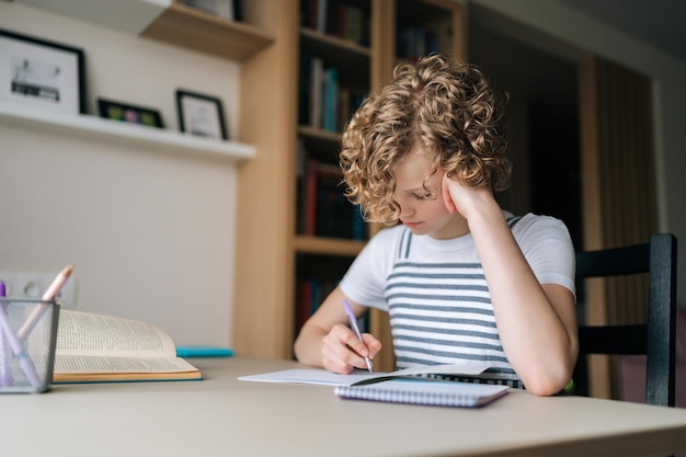 Retrato de uma colegial pensativa escrevendo no livro de exercícios fazendo lição de casa sentada na mesa perto da janela em casa