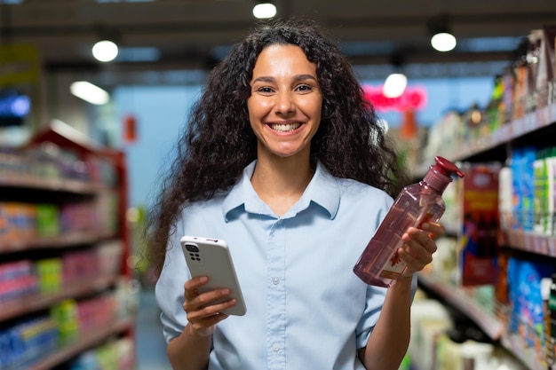 Retrato de uma compradora em um supermercado uma mulher hispânica escolhe shampoo para o cabelo e gel de banho lê críticas na Internet sorri e usa um aplicativo em seu telefone