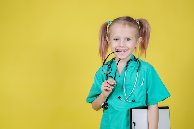 Foto retrato de uma criança caucasiana sorridente, vestida com um jaleco verde de médicos e um tablet isolado em amarelo na mão.