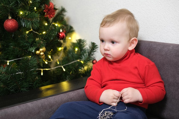 Retrato de uma criança no fundo de uma árvore de Natal. Menino brinca com um presente.