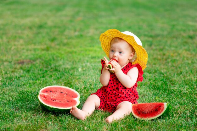 Retrato de uma criança no verão na grama verde comendo uma melancia em um espaço de chapéu amarelo para texto