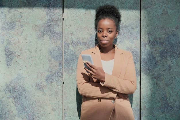 Retrato de uma empresária africana de terno, olhando para a câmera e usando seu telefone celular ao ar livre