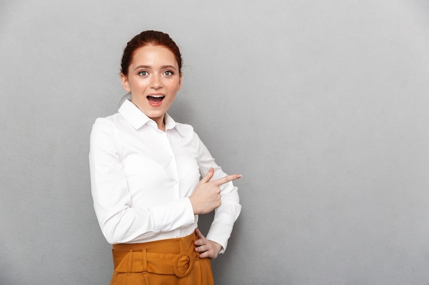 Retrato de uma empresária ruiva satisfeita de 20 anos com roupa formal, sorrindo e posando em um escritório isolado sobre o cinza