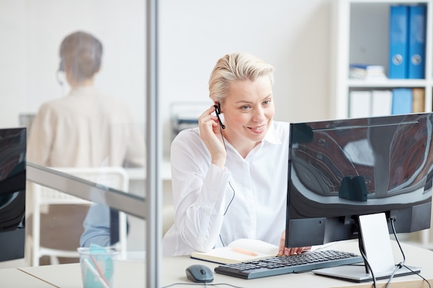 Retrato de uma empresária sorridente, falando ao microfone enquanto usa o computador no interior do escritório, conceito de suporte ao cliente