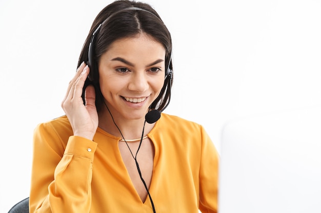 Retrato de uma encantadora assistente de linha direta usando fone de ouvido com microfone, falando com o cliente por telefone enquanto trabalha no escritório