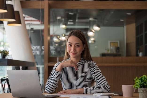 Retrato de uma encantadora empresária asiática sentada trabalhando no laptop no escritório olhando para a câmera