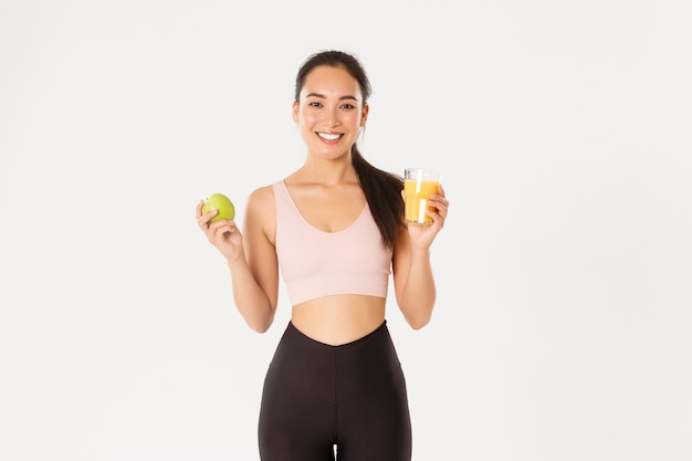 Retrato de uma esportista asiática bonita saudável e em forma segurando suco de laranja e maçã, café da manhã antes do treino