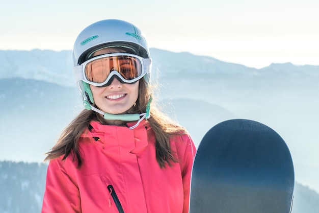 Retrato de uma esportista usando capacete e máscara com snowboard na mão, olhando para a câmera, sorrindo, aproveitando o dia ensolarado e gelado