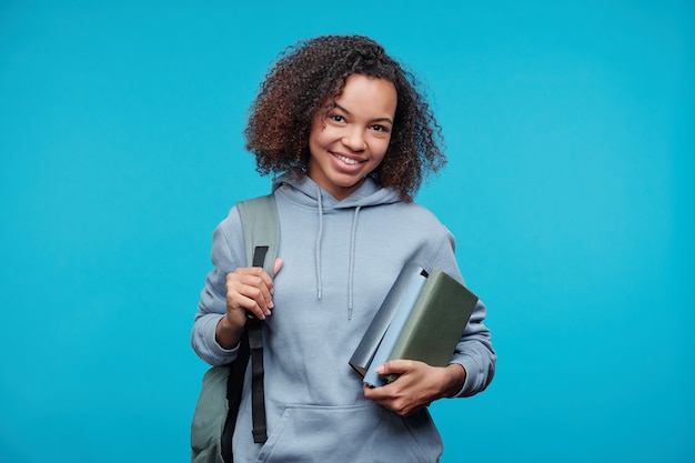 Retrato de uma estudante afro-americana positiva com capuz segurando uma pilha de livros contra um fundo azul
