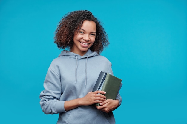 Retrato de uma estudante afro-americana sorridente com um capuz segurando livros e em pé contra um fundo azul