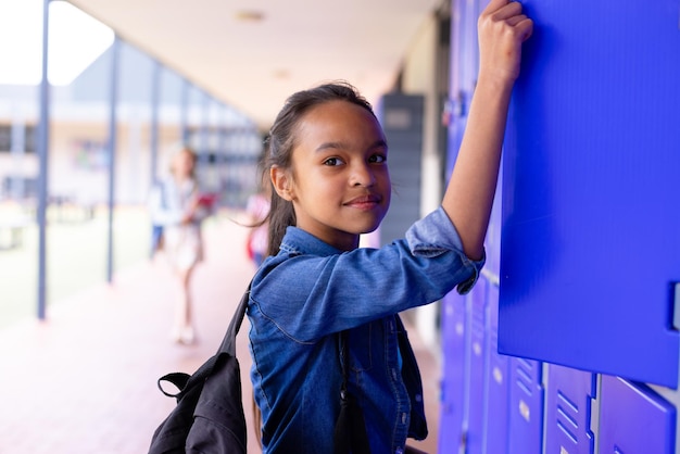 Foto retrato de uma estudante biracial feliz de pé ao lado de armários no corredor da escola