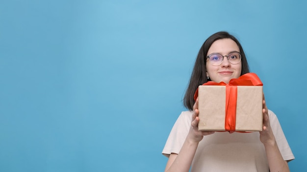 Retrato de uma estudante feliz sorridente com óculos e uma blusa bege segurando uma grande caixa de presente com um laço vermelho