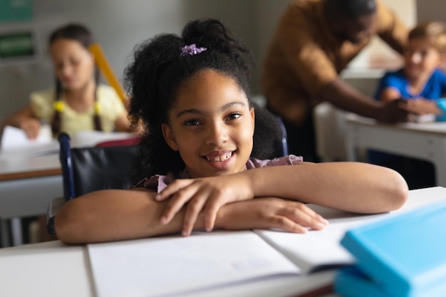 Retrato de uma estudante primária biracial sorridente sentada em cadeira de rodas em uma mesa na sala de aula