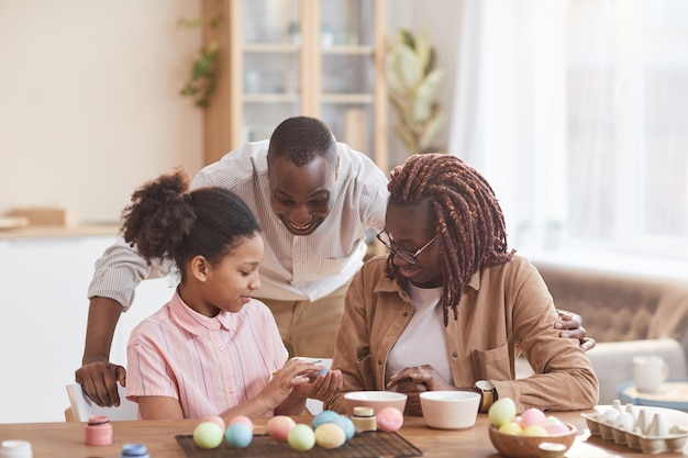 Retrato de uma família afro-americana adorável pintando ovos de Páscoa juntos, sentados à mesa de madeira em um interior aconchegante e apreciando a arte DIY, copie o espaço